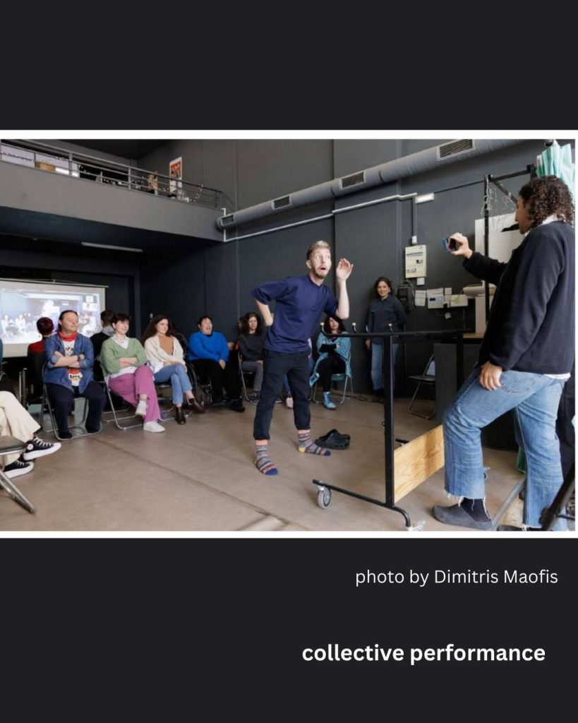 Φωτογραφία σε μαύρο πλαίσιο, από χώρο performance/στούντιο με ψηλή οροφή, εκτεθειμένους σωλήνες και εσωτερικό μπαλκόνι στο βάθος. Στο κέντρο, ένας άντρας με ξανθά μαλλιά, μπλε μπλούζα και μαύρο παντελόνι, ξυπόλητος με κάλτσες σε διαφορετικά χρώματα, βρίσκεται σε δυναμική στάση με το ένα χέρι σηκωμένο και έκφραση έκπληξης στο πρόσωπο — φαίνεται να αυτοσχεδιάζει ή να παίζει θέατρο. Δεξιά, μια γυναίκα με σγουρά μαλλιά, σκούρα ζακέτα και τζιν, στέκεται πάνω σε ξύλινη/μεταλλική κατασκευή και τον φωτογραφίζει με το κινητό της τηλέφωνο. Αριστερά, μια ομάδα θεατών κάθεται σε καρέκλες παρακολουθώντας τη σκηνή — φαίνονται περίπου δέκα άτομα διαφορετικών ηλικιών, με casual ρούχα σε ποικίλα χρώματα. Στο βάθος αριστερά προβάλλεται μια οθόνη με εικόνα. Κάτω από τη φωτογραφία, σε μαύρο φόντο, υπάρχει η λευκή γραφή «photo by Dimitris Maofis» και πιο κάτω, με έντονα γράμματα, η φράση «collective performance».