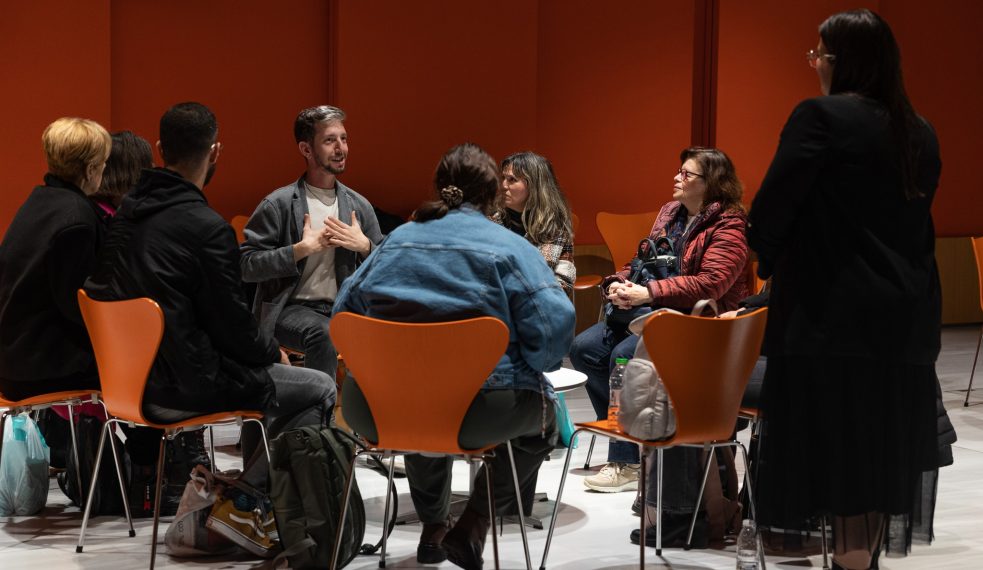 Ομάδα ανθρώπων κάθεται σε κύκλο σε έναν φωτεινό χώρο και συμμετέχει σε συζήτηση. Οι συμμετέχοντες έχουν οπτική επαφή μεταξύ τους, ενώ ένα άτομο μιλάει και χρησιμοποιεί τα χέρια του για να εκφραστεί. Η κυκλική διάταξη διευκολύνει την οπτική επικοινωνία και τη συμμετοχή όλων.